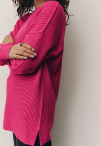 Pulover tricotat fucsia cu croială lejeră, fante laterale și manșete cu model din rib. Textura este moale și texturată, având guler rotund.
