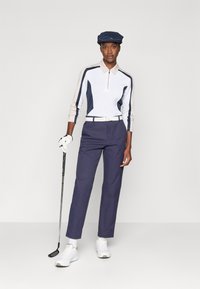 Golf-Outfit mit einem weißen und marineblauen Langarm-Poloshirt, marineblauen Hosen, weißem Gürtel und weißen Schuhen. Zubehör umfasst eine marineblaue Kappe und einen Golfschläger-Handschuh.
