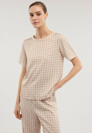 Frau trägt beige-weiß kariertes kurzärmeliges Oberteil und passende Hose, steht mit einer Hand in der Tasche vor einfarbigem Hintergrund.