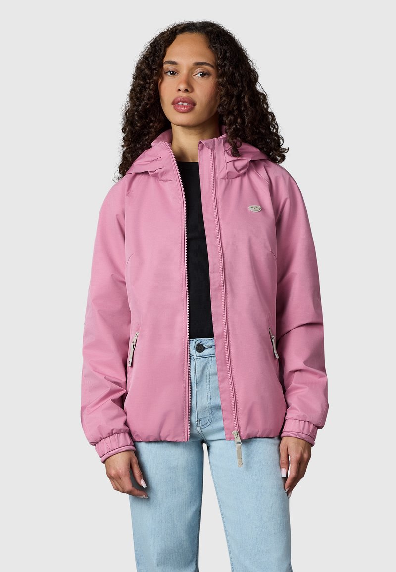 Vrouw met krullend haar die een roze jas met rits, een zwart topje en lichtblauwe spijkerbroek draagt tegen een effen lichtgrijze achtergrond.