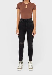 Mujer con una blusa marrón de manga corta y escote en clave, jeans skinny rasgados de cintura alta negros y zapatillas blancas y verdes, de pie contra un fondo blanco.