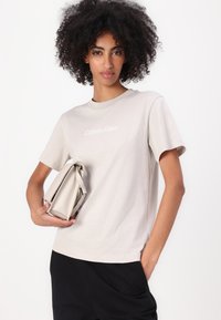T-shirt din bumbac gri deschis, cu mâneci scurte și guler rotund, având un logo alb "Calvin Klein". Modelul ține o geantă de culoare deschisă.