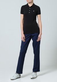 Schwarzes Polohemd mit Kragen und drei Knöpfen, kombiniert mit marineblauer Hose und weißen Sportschuhen. Der Stoff wirkt glatt und anliegend.