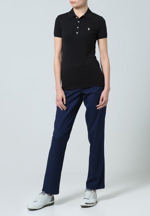 Černé polokošile s límečkem a třemi knoflíky, doplněné tmavě modrými kalhotami a bílými sportovními botami. Materiál působí hladce a přiléhavě.