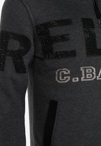 Grauer Pullover mit großen schwarzen Strassbuchstaben und kontrastierendem Drucktext. Enthält eine Fronttasche und einen Reißverschluss am Hals.