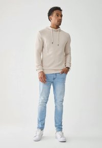 Mladý muž stojí, oblečený v béžové mikině, světle modrých džínách a bílých teniskách, na jednobarevném světlém pozadí.