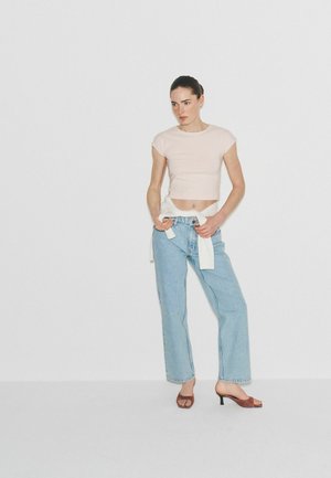 Kvinde iført en lys cropped t-shirt, lyseblå lige jeans, bundet hvid sweatshirt rundt om taljen og brune hælede sandaler, der står mod en hvid væg.