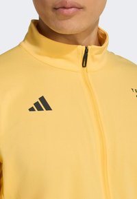 Gele sportjas met een hoge kraag en rits aan de voorkant. Voorzien van een zwart Adidas-logo en tekst op de borst. Gladde stoftextuur.