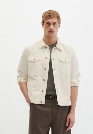 Blondyn z poważnym wyrazem twarzy, ubrany w kremową zapinaną na guziki kurtkę na szarej koszuli i ciemnobrązowych spodniach, z rękami w kieszeniach.