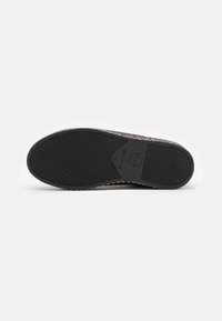 Kötött slip-on cipő talpa fekete textúrált gumival, a boltozat közelében logóval, fehér háttéren.