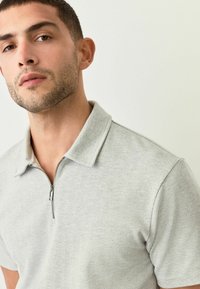 Polo de color gris claro con patrón de espiga, con cuello con cremallera, mangas cortas y tejido texturizado. Vista cercana del escote y la parte superior del torso.
