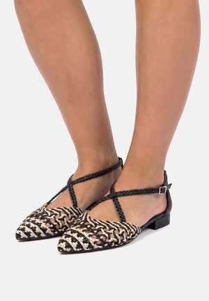 Pies con bailarinas tejidas en negro y beige de punta afilada, con correas trenzadas en el tobillo y pequeños tacones cuadrados sobre un fondo claro.