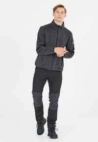 Chaqueta de cremallera gris oscuro con un cuello alto y dos bolsillos laterales; se lleva con pantalones negros que tienen parches grises en las rodillas. Botas negras completan el atuendo.