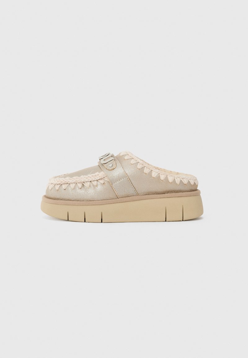 Beige slip-on klipp med metallisk glans, med teksturert søm og en frontstropp med en sølvspenne. Kraftig, teksturert såle.