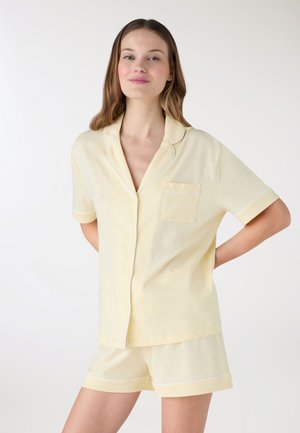 Giovane donna che indossa una camicia giallo chiaro a maniche corte con bottoni e pantaloncini coordinati, in piedi con le mani dietro la schiena.