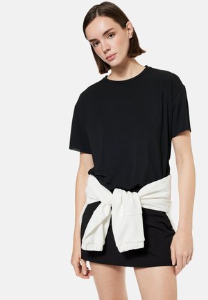 Giovane donna che indossa un vestito maglietta nero oversize con felpa bianca legata in vita, in piedi davanti a uno sfondo chiaro e uniforme.