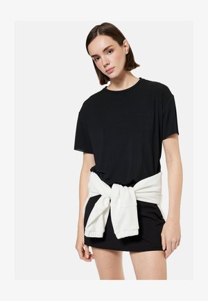 Młoda kobieta ubrana w czarną, oversize sukienkę-t-shirt, z białą bluzą zawiązaną w talii, stoi na tle jednolitego jasnego tła.