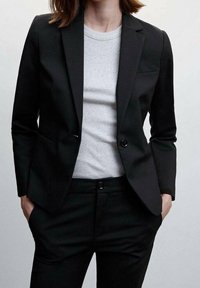 Zwarte op maat gemaakte blazer met een slim fit, enkele knoopsluiting en geknipte revers, gedragen over een lichtgrijze fitted blouse en bijpassende zwarte broek.