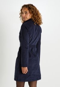 Femme aux cheveux bouclés portant une robe longue à manches longues bleu marine ceinturée, regardant par-dessus son épaule sur un fond blanc.