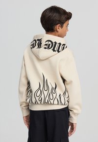 Beżowa bluza z kapturem z dużym czarnym napisem na kapturze i czarnymi wzorami płomieni na dolnym wykończeniu. Miękki materiał o luźnym kroju.