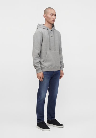 Homme debout en sweat à capuche gris, jean bleu et baskets noires, sur un fond uni gris clair, légèrement tourné vers la droite.