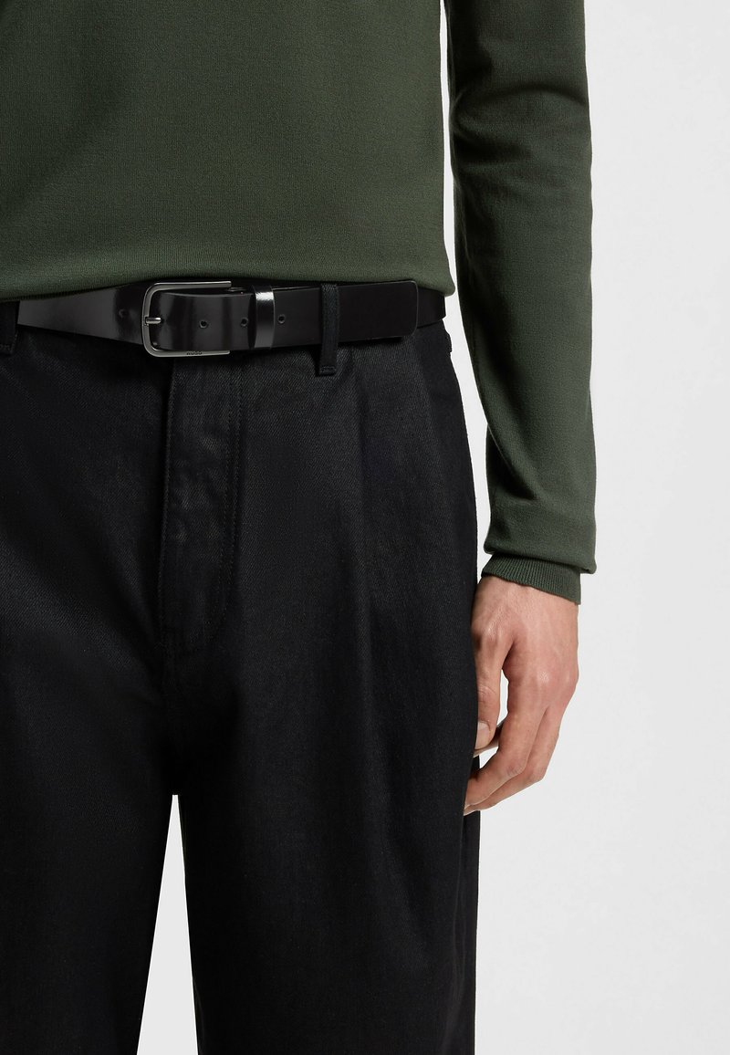 Cintura in pelle nera con fibbia rettangolare in argento, indossata sopra pantaloni in tessuto plissettato scuri, abbinata a una camicia verde a maniche lunghe.