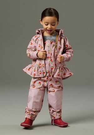 Taapero, jolla on päällään vaaleanpunainen sadeasu, jossa on mansikkakuvio, vetää vetoketjua alas, punaiset kumisaappaat päällä, yksiväristä taustaa vasten.