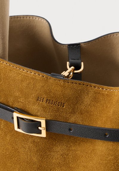 Brun ruskind tote taske med en sort læderrem og hardware i guldfarve. "BEE PROJECTS" præget på ruskindet. Enkel, ustruktureret design.