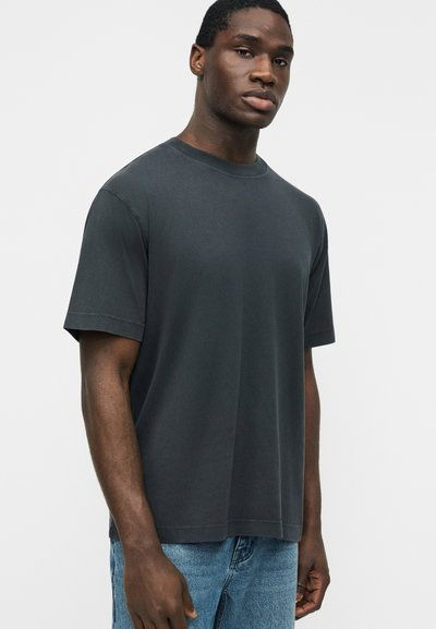 Νεαρός άνδρας με σκουρόχρωμο δέρμα που φοράει απλό κοντομάνικο μπλουζάκι σε σκούρο γκρι και μπλε τζιν παντελόνι, στέκεται μπροστά σε ανοιχτό γκρι φόντο.