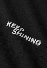 Schwarzer Stoff mit weißem, besticktem Text "KEEP SHINING" in einer fetten, leicht strukturierten Schrift. Einfaches und klares Design.