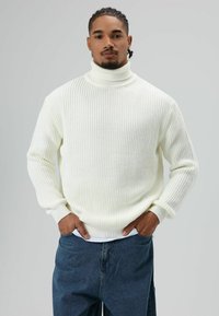 Man met gevlochten haar, gekleed in een witte coltrui en blauwe spijkerbroek, staand met handen in de zakken tegen een grijze achtergrond.