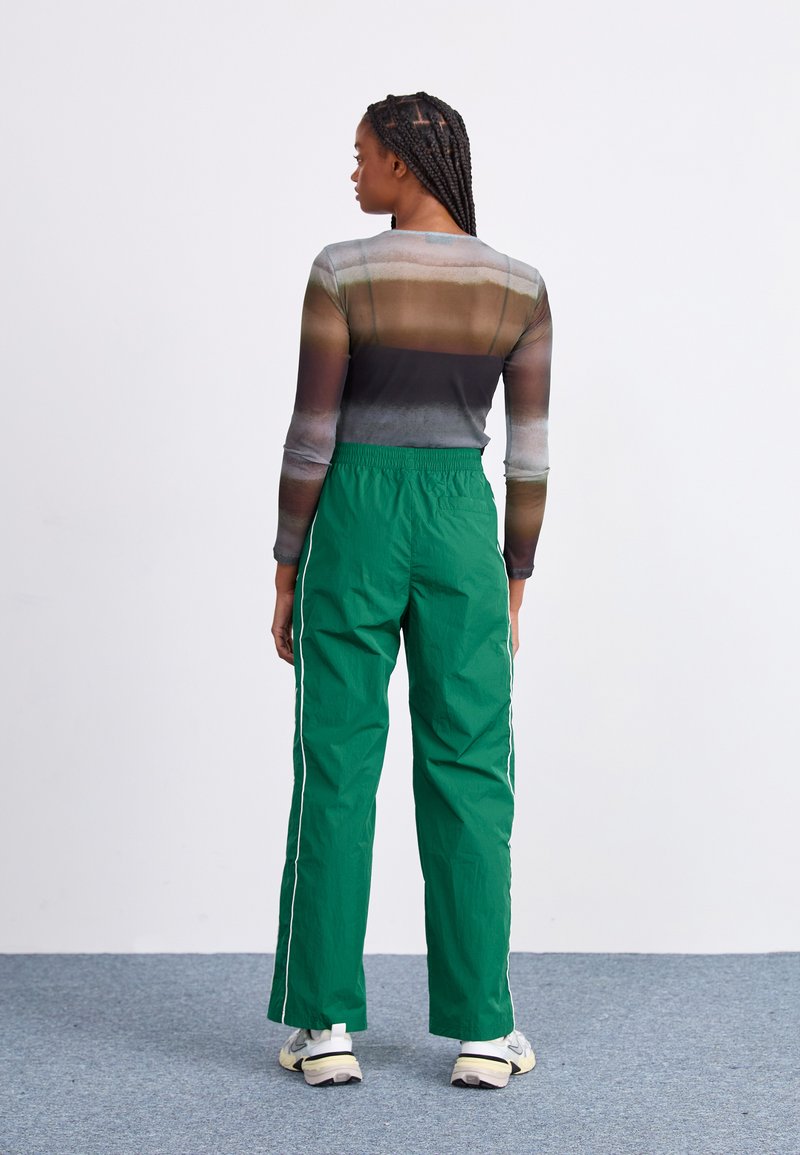 Groene lichtgewicht broek met witte zijstrepen, gecombineerd met een semi-doorzichtige, meerkleurige lange mouw top, gezien van achter.