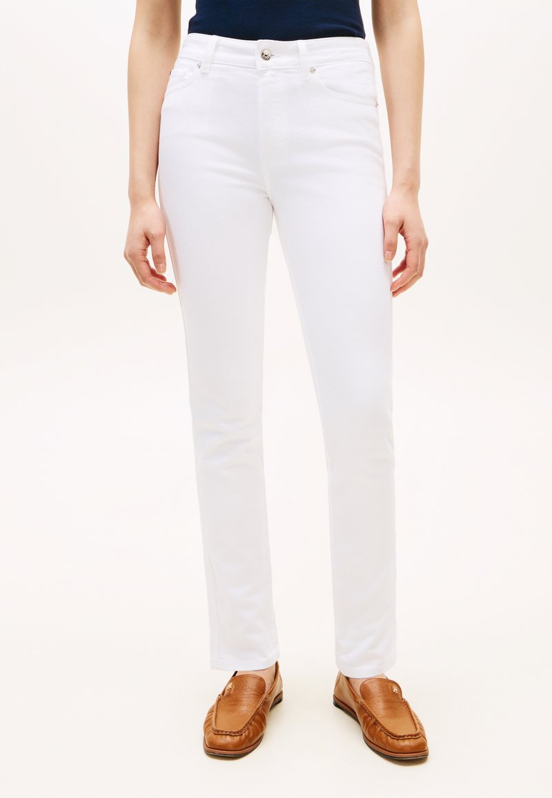 Γυναίκα που φοράει λευκό παντελόνι στενής γραμμής και καφέ δερμάτινα μοκασίνια, στέκεται μπροστά σε απλό φωτεινό φόντο.