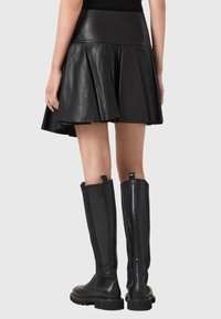 Γυναίκα που φοράει μαύρη δερμάτινη φούστα με φουσκωτή γραμμή και μαύρες δερμάτινες μπότες μέχρι το γόνατο, εμφανίζεται από πίσω μπροστά σε ένα απλό φόντο.