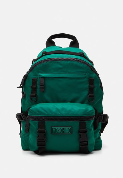 Zielony nylonowy plecak z czarnymi akcentami, wyposażony w liczne zasuwane komory, regulowane paski oraz przednią kieszeń oznaczoną napisem "MOSCHINO."