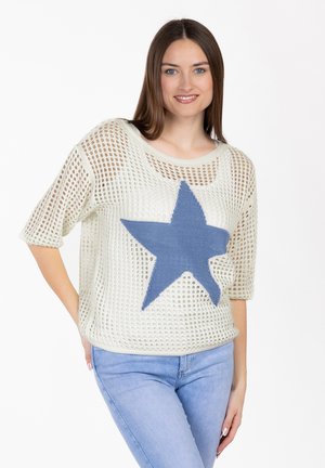 Młoda kobieta uśmiechająca się, ubrana w biały sweter z siateczki z dużą niebieską gwiazdą na przodzie i jasnoniebieskie dżinsy, stojąca na białym tle.