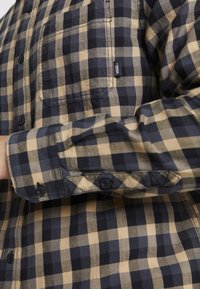 Camicia buttons-up con un motivo a scacchi nero e beige, realizzata in tessuto morbido, dotata di taschino sul petto e bottoni neri.