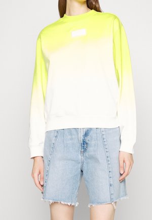 Femme portant un sweat à manches longues avec un dégradé jaune néon vers blanc et un short en denim bleu clair taille haute avec des ourlets effilochés.