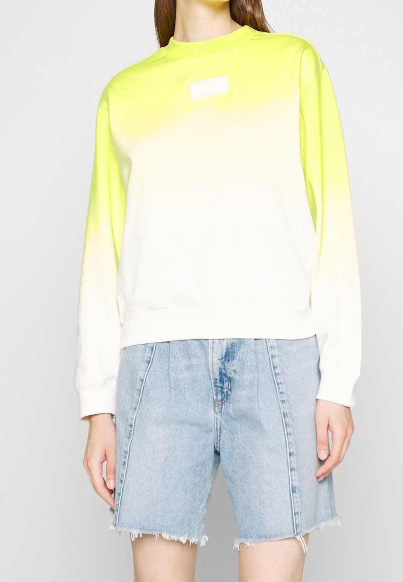Femme portant un sweat à manches longues avec un dégradé jaune néon vers blanc et un short en denim bleu clair taille haute avec des ourlets effilochés.