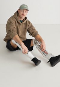 Pierna prostética blanca con un diseño transparente, llevada por una persona con una sudadera marrón y pantalones cortos negros, sentada sobre una superficie clara.