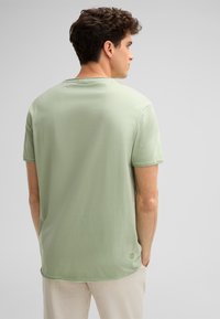 Mintgrünes Baumwoll-T-Shirt mit rundem Halsausschnitt und kurzen Ärmeln, das einen etwas längeren hinteren Saum und ein dezentes Logo-Detail an der Seite aufweist.