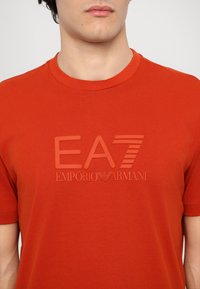 Red cotton t-shirt featuring a raised rubberized logo with "EA7" and "EMPORIO ARMANI" text. Short sleeves and a classic crew neck design.