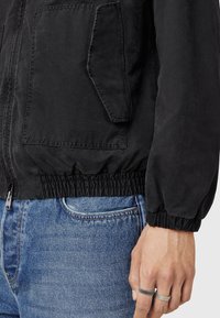 Sort jakke med elastiske manchetter og kant, med lynlås og en fremadrettet lomme. Bæres over blå denim jeans.