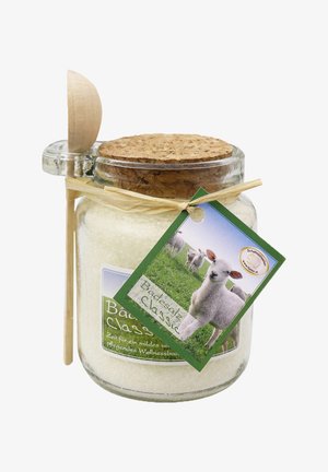 Ein klarer Glasbehälter mit Badesalz, einem Korkdeckel und einem Holzlöffel. Das grüne Etikett zeigt Schafe auf Gras und kennzeichnet den Produktnamen "Badesalz Classic."