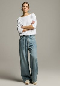 Biała, luźna, długozasięgowa bluzka zestawiona z szerokimi jasnoniebieskimi dżinsami. Z wyróżniającym się sznurkiem w talii i kieszeniami.