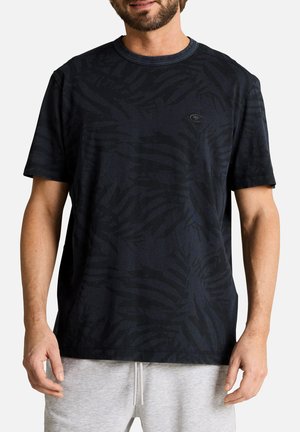 Mann trägt ein schwarzes kurzärmeliges T-Shirt mit dezentem Blattmuster und hellgraue Jogginghose, steht vor weißem Hintergrund.