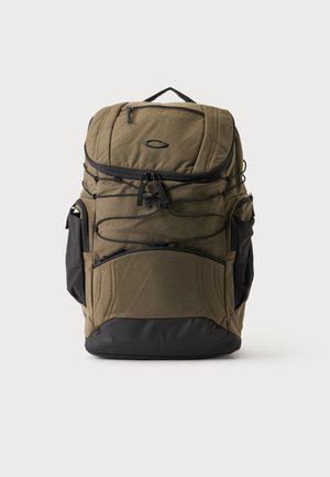 Sac à dos vert olive avec plusieurs compartiments zippés, fond noir, poches latérales, cordons élastiques et une poignée de transport supérieure sur fond blanc.