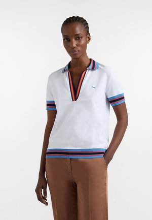 Donna con i capelli intrecciati che indossa una polo bianca a maniche corte con colletto, polsini e orlo a righe blu, rosse e nere, abbinata a pantaloni marroni.