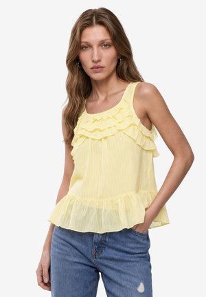 Donna che indossa un top giallo senza maniche con volant e jeans blu, con una mano in tasca, su uno sfondo bianco uniforme.
