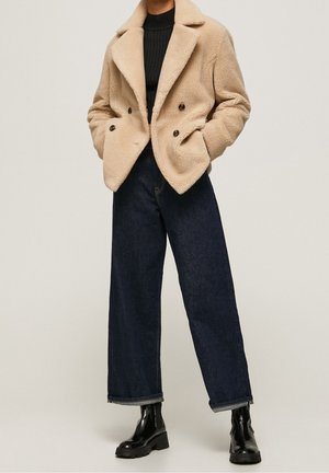 Osoba ubrana w beżową dwurzędową kurtkę z sztucznego futra, czarny golf, ciemne szerokie jeansy oraz czarne masywne botki za kostkę.